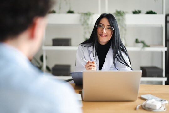 A doctor in a white coat smiles while holding a stylus, looking at a patient across a desk with a laptop and tablet.