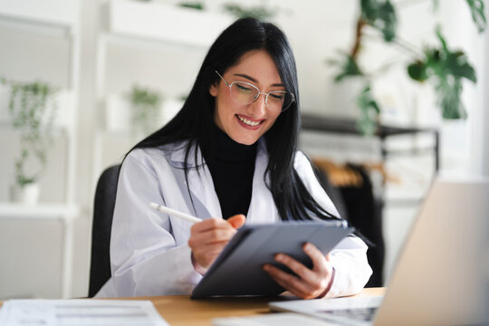 A smiling woman in a lab coat uses a stylus to write on a tablet computer while working at her desk.