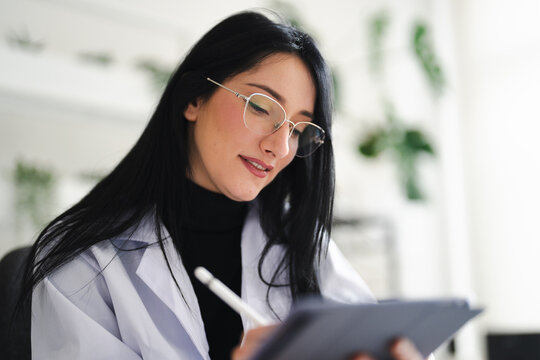 A woman in a lab coat and glasses uses a stylus to write on a tablet, focusing on her work.
