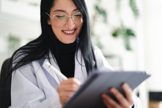 A smiling woman in a lab coat uses a stylus to write on a tablet, engaged in her work.