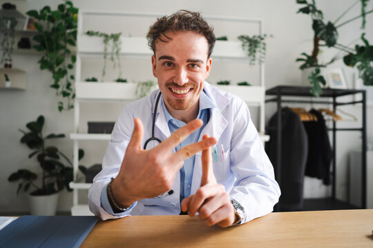 A smiling doctor in a white coat gestures with his hands while sitting at a desk in a bright, plant-filled office.