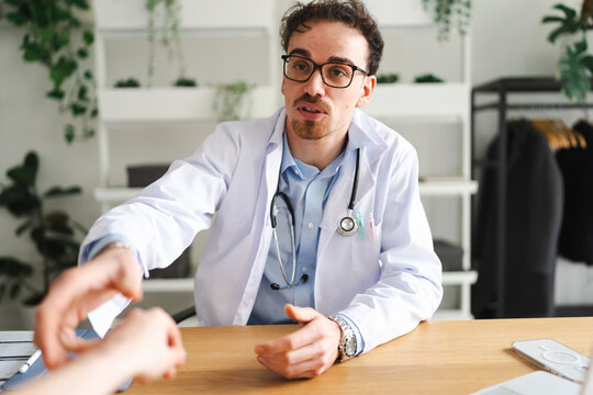 A doctor in a white coat and stethoscope is talking to a patient and reaching out their hand across a wooden desk in a modern office.