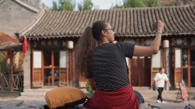 Woman sitting stone wall photographing heritage village with phone polkadot top, red jacket tied at waist, yellow cushion beside traditional wooden doors, tiled roofs and lanterns in background casual