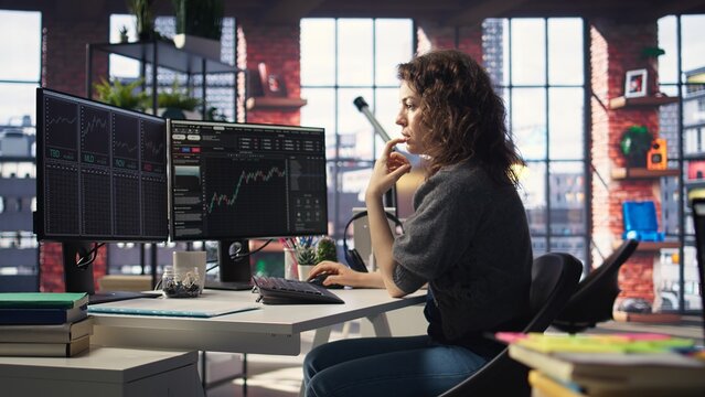 Female day trader examines stock market graphs on dual monitors, tracking financial growth to share prices. Trading interface provides real time data for buying and selling shares. Camera A.