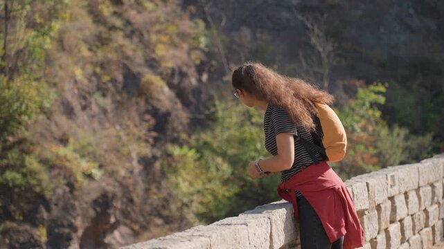 Young woman on lookout photographing canyon with smartphone, leaning on stone wall, orange backpack, focused on angle and light while scouting scenic composition and documenting travel memories.