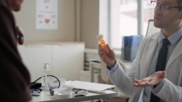 Urologist presenting medicine bottle and discussing therapy options with adult patient during renal consultation in modern office setting, eyelevel medium shot