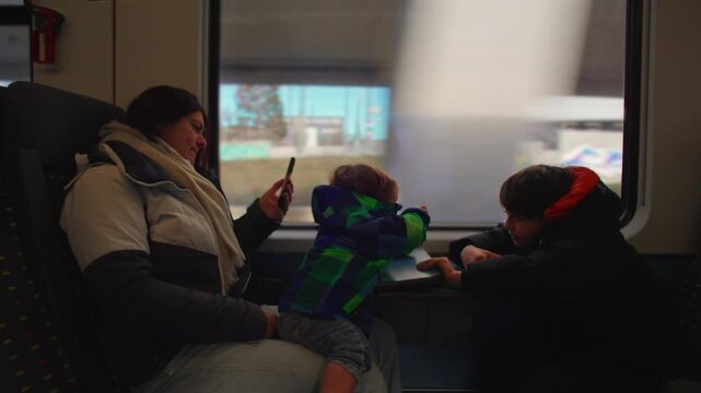 Mother traveling by train with two sons, family seated around cabin table as children watch scenery through window during relaxed rail journey