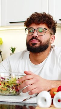 Arabian man stares at salad bowl in kitchen feeling exhausted from strict keto diet and healthy eating at home. Young guy loses appetite, wants comfort food but keeps trying weight loss discipline