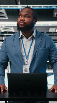 Vertical video Server farm programmer at workplace desk using laptop to optimize electronics. African american man in data center using notebook to monitor system performance, identifying bottlenecks