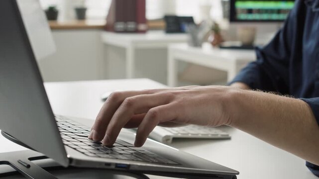 Rack focus shot of male programmers hands typing on wireless computer keyboard then switching to laptop while writing script on devices at desk in office