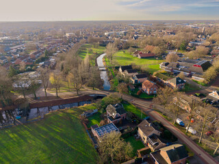 Fototapeta premium Aerial view of a tranquil European town with a winding canal, green parks, and brick houses under a soft morning sun. A peaceful suburban landscape with traditional architecture and nature.