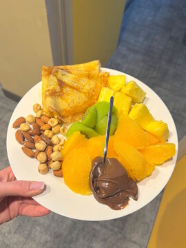 hand holding white plate with food of sliced ​​fruits chocolate and pancakes in room