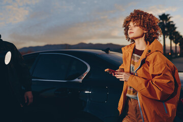 A woman in a rainbow sweater and orange jacket smiles while holding a smartphone near a black car...