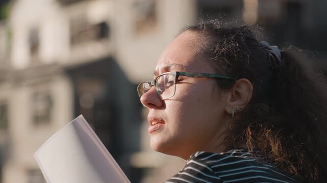 Multiracial woman aspiring architect observing skyline notebook raised, glasses catching sunlight, urban building backdrop, profile shots, pen poised, studying form and proportion, reflective mood