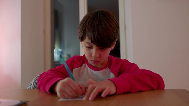 Young boy sitting at table carefully writing on small cards with colored pencil focusing on homework task and developing concentration and fine motor skills