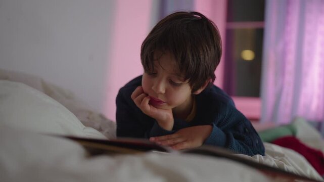 Young child lying on bed reading book at night near window, thoughtful expression and quiet focus during bedtime story moment at home interior