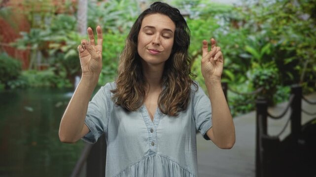 Woman wearing denim dress makes air quotes with hands at green forest walkway near water pond; skepticism.