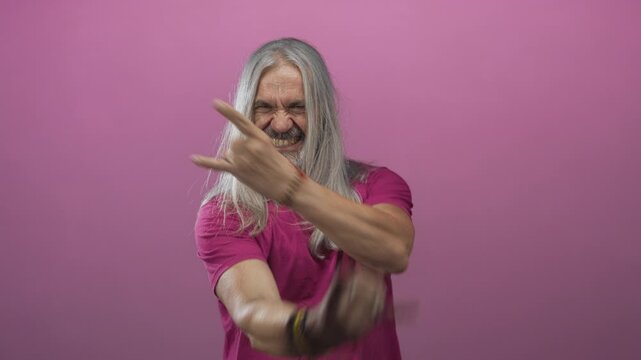 Man middle age with hoary long hair grinning while showing rock horns hand gesture in pink studio with bracelets and rings visible; rebellion attitude energy.