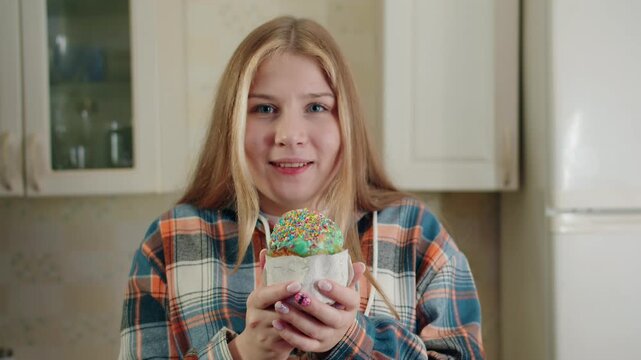 Caucasian smiling woman with flannel shirt holding easter decorated cupcake with green glaze in colored sprinkles in illuminated kitchen. Her eyes closed while inhaling sweet aroma. Look at camera
