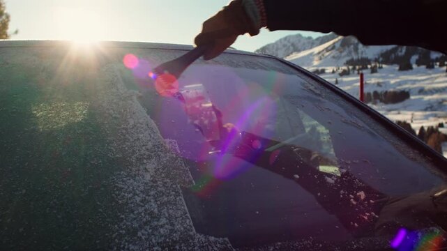 Gloved hand clearing frozen windshield with ice scraper under bright winter sun, snow covered hills and blue sky behind parked car