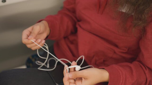 Close up careful hands separating knotted earbuds on lap, student methodically teases cords apart above phone, focused preparation before study with steady dexterous movements and warm interior light.