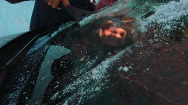 Hand scraping ice from car windshield with reflection visible in frozen glass, winter morning scene preparing vehicle for cold weather drive