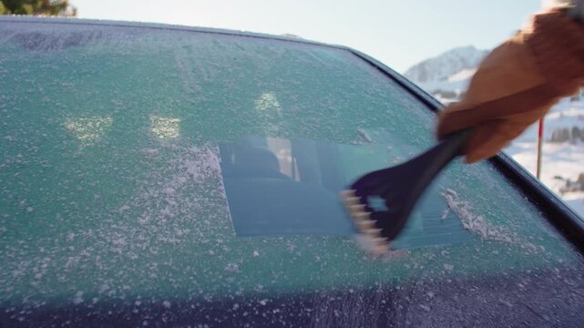 Hand in winter glove scraping ice from car windshield in snowy mountain setting, close view of frost removal during cold morning commute in alpine landscape