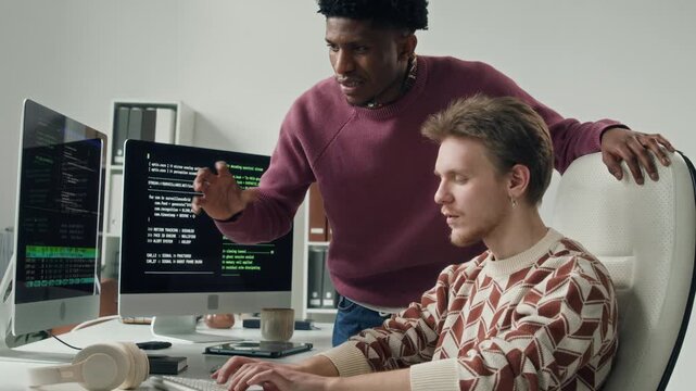 Side view of young adult Caucasian male software engineer typing on computer keyboard while pair programming with Black male colleague reviewing code on screen at office workstation