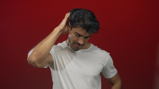 Man in white t-shirt scratching his head in a red studio setting while looking down with puzzled look; confusion doubt uncertainty hesitation.