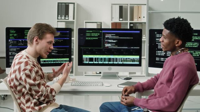 Side view of professional male software developer discussing code optimisation with Black male coworker sitting at workstation with multiple screens displaying script then cheerfully clasping hands