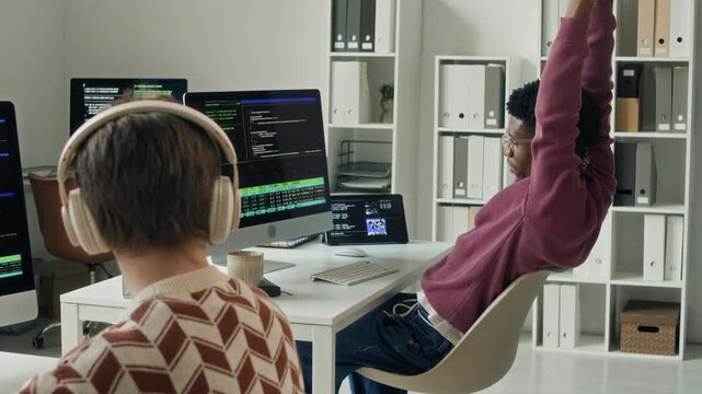 Side view of young adult African American male developer stretching out arms after long computer work at office desk then consulting colleague on work task during collaborative work in open space