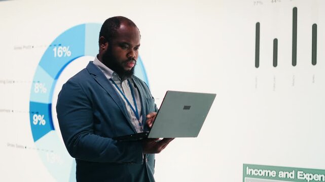 Manager analyzes revenue, expenses and net profit displayed on presentation wall using laptop. African american man uses notebook to review quarterly KPIs results, camera B