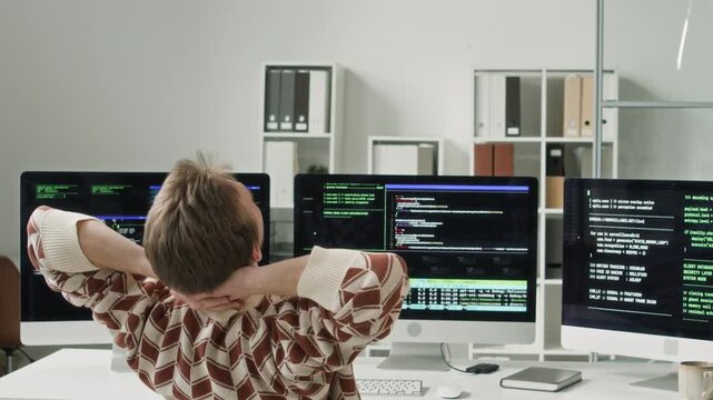 Rear view of young adult male IT engineer stretching out arms and back while sitting at workstation with multiple computer monitors displaying code in modern office