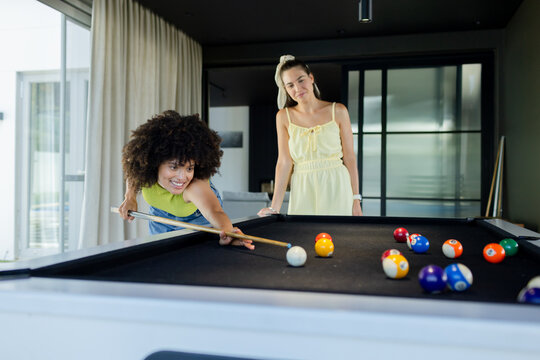 Female friends playing pool in game room, leaning over black-felt table lining up cue ball