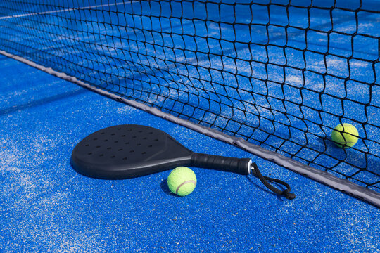 Black padel racket lying on blue synthetic court with sand granules, two neon balls near net