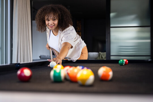 African American adult woman in casual clothing leaning over black-felt pool table aiming cue ball