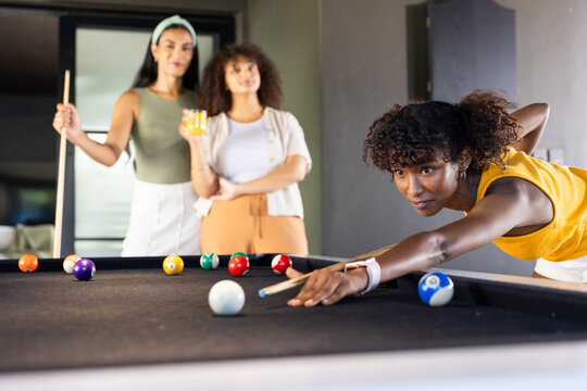 African woman in yellow top leaning over pool table at home, lining up shot, copy space