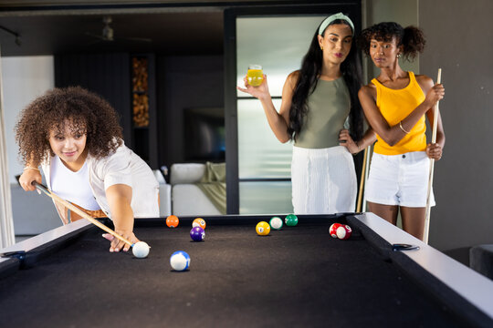 African American women leaning over black felt pool table lining up shots with cues, glass