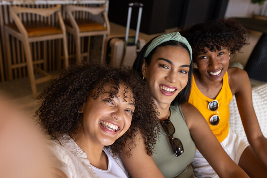 Female friends posing for selfie in lounge wearing sleeveless tops and sunglasses with suitcase