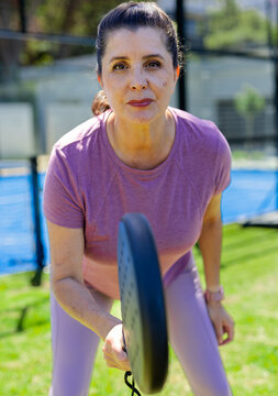 Mature woman leaning holding round paddle on fenced blue court wearing purple sportswear and watch