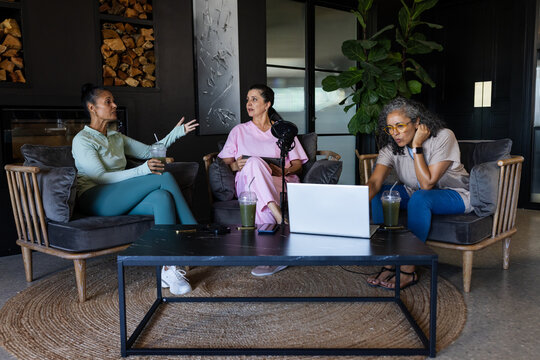 Diverse female colleagues wearing loungewear recording in studio near mic, laptop and green drinks