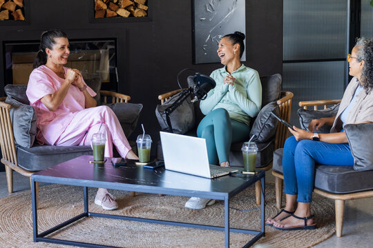 Diverse female collaborators chatting in lounge studio with microphone, laptops, tablet, smoothies