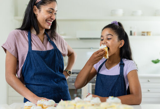 African American mom and daughter baking, tasting cupcakes in kitchen in denim aprons, copy space
