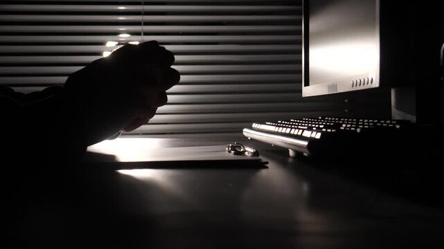 Hands in silhouette crumpling a sheet of paper at a computer workstation, depicting concepts of stress, frustration, failure, and workplace anger in a dim office environment