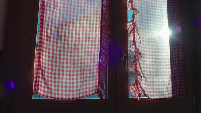 Pair of red and white checkered curtains covering wooden kitchen window with sunlight streaming through fabric, nostalgic country home interior detail