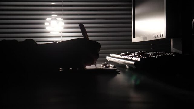 Person's hand making notes with a pen on paper at a dark desk, illuminated by a computer monitor and light from venetian blinds, symbolizing hard work and dedication