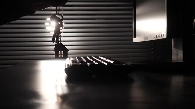 Hand silhouette holding a house key ring with a small house charm, contrasting against a bright window and a dark office desk with a computer monitor and keyboard