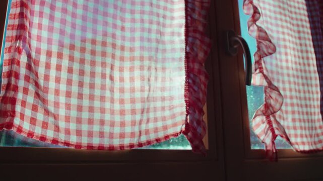 Red and white checkered kitchen curtains glowing in morning sunlight through window, rustic home interior detail with fabric texture and gentle movement