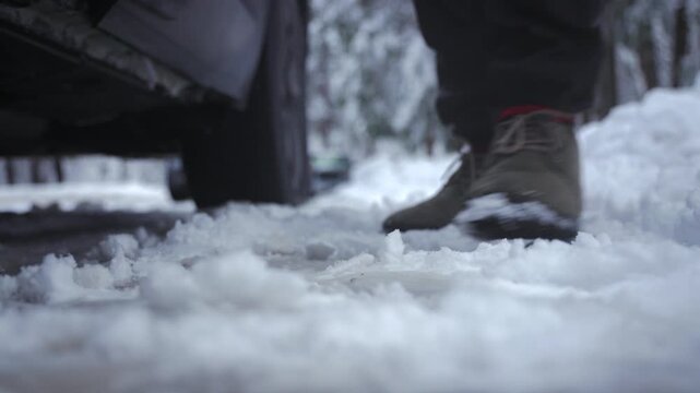 Closeup boot stepping on snow beside parked car, pine forest backdrop, tire and icy road visible, red sock and tied laces, hiker exiting vehicle, crunching footprint in frost, cold blue