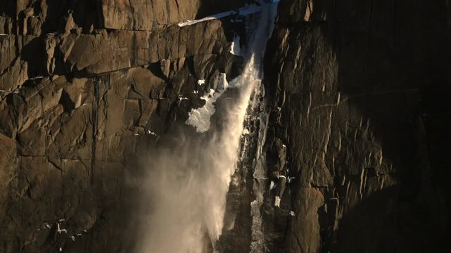 Golden light on granite Yosemite waterfall cascading across cliff face, winter snow patches clinging to ledges, heavy mist and roaring spray, warm highlights and deep shadow contrast, cinematic